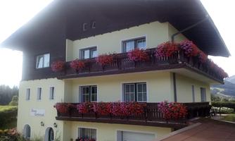 A charming building with a balcony full of blooming flowers. The façade is bright and friendly, surrounded by beautiful scenery.