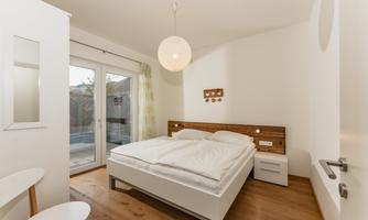 A modern bedroom with a double bed and wooden details. Generous windows provide plenty of daylight.