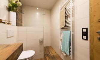 A modern bathroom with a white tiled wall and wooden details. It is bright and friendly, with a towel holder and a small sink.
