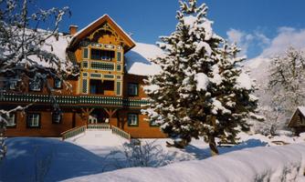 A charming wooden house in the snow, surrounded by snow-covered trees. The sky is clear and blue, creating a serene winter atmosphere.