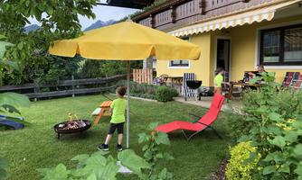 A cozy garden with a yellow sunshade and a grill. Two children are playing in the green next to a red deck chair.