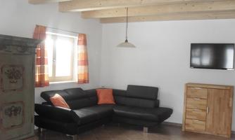 A modern living room with a black couch and a wooden table. On the wall, there is a television and a colored curtain lets light in.