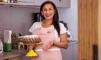 A smiling woman stands in a modern kitchen holding a cake on a yellow plate. She is wearing an apron and appears to be happy about her baked goods.