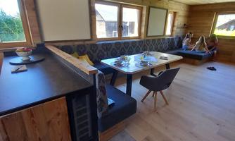 A bright living room with wooden ceilings and flooring. There is a large table and a cozy seating area where several people are sitting.