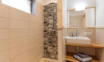 A modern bathroom with a stone shower and bright tiles. The washing area features a sink and wooden shelves.