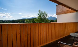 A balcony with a wooden wall and a beautiful view of green trees and mountains. The sky is clear and blue.
