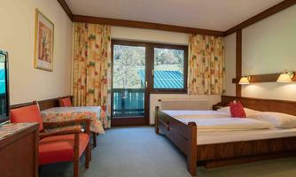 A simple hotel room with two beds, a table and chairs. The room has large windows overlooking a balcony and colorful curtains.