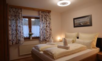 A cozy bedroom with a double bed and bright pillows. Large windows with curtains and soft lighting create an inviting atmosphere.