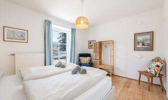 A bright bedroom with two beds and a cozy armchair. Large windows and warm wooden paneling create an inviting atmosphere.