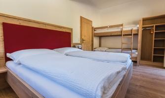 A cozy room with two beds and a bunk bed in the background. The walls are light and the floor is wooden.