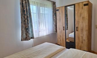 A bright bedroom with two beds and a wooden wardrobe. Transparent curtains hang in front of the window.