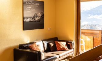A cozy living room with a dark sofa and decorative cushions. A picture of mountains hangs on the wall, and a window shows a view of nature.