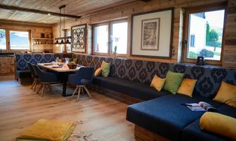 A cozy living area with a large sofa and a dining table. The walls are made of wood, and large windows let in plenty of light.