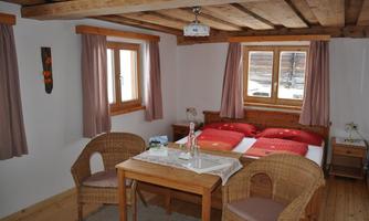 A cozy room with a double bed and a small table. It has wooden walls, large windows, and is bright and inviting.