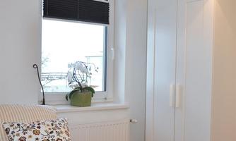 A bright bedroom with a white wardrobe and a window. On the bed lies a pillow with floral patterns.
