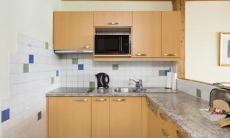 A modern kitchen unit with wooden cabinets and a marble countertop. The equipment includes a microwave, a kettle, and a refrigerator.