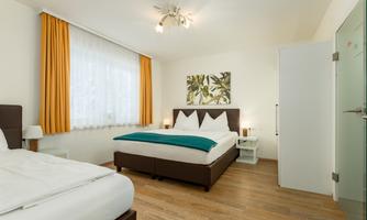 A modern bedroom with two beds and wooden flooring. The windows are adorned with light curtains, and there is a stylish wall decoration.