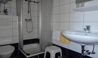 A simple bathroom with a shower, a sink, and a toilet. The walls are tiled and there is a white stool.