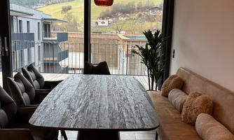 A modern dining room with a large table and cozy chairs. Through the windows, there is a view of the green landscape.