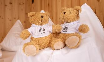 Two cuddly bears are sitting on a bed. Both are wearing T-shirts with prints.