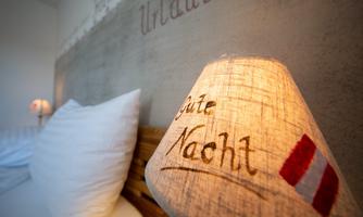 A cozy bedroom with a wooden frame bed and large white pillows. A lamp with the inscription "Good Night" creates an inviting atmosphere.