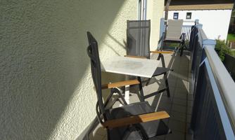 A balcony with a small table and two chairs. The sun is shining, and the surroundings are quiet.