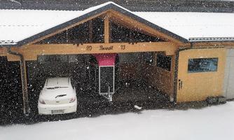 A covered parking space with a white car and a pink wooden cover. It is snowing heavily in the background.