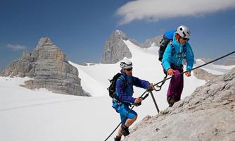 Two climbers are climbing a rocky wall with snow-covered peaks in the background. The sky is clear and blue.