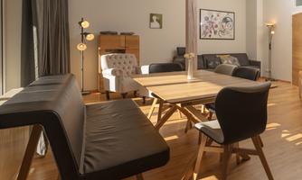 A modern living room with a wooden table and various seating options. Soft light filters through the windows, creating an inviting atmosphere.