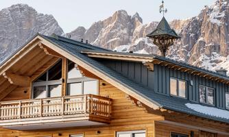 A modern wooden house with a balcony and a large, pointed roof. In the background, majestic mountains can be seen.