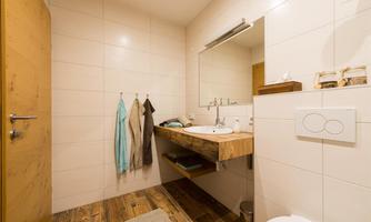 A modern bathroom with a wooden vanity and a large mirror. Some towels are hanging on the wall, and the floor is tiled with wooden tiles.