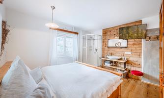 A modern bedroom with a large bed and wooden elements. Next to the bed, there is a bright bathroom and a television on the wall.