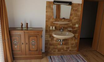 A simple bathroom with a sink and mirror on a tiled wall. Next to the sink is a wooden cabinet.