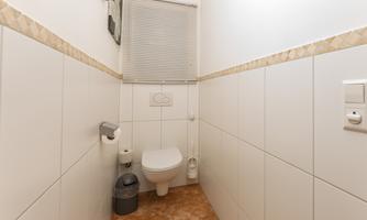 A simple bathroom with white tiles and a modern toilet. The window is fitted with a blind, and a trash can is visible.