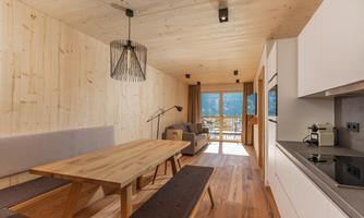 A modern living and dining room with wooden walls and a large dining table. Large windows provide a view outside and plenty of natural light.