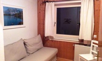 A small room with a sofa and a white chair. The walls are wooden and a window lets in daylight.