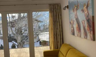 A bright and modern living room with a yellow sofa and a small table. Large windows offer a view of nature.