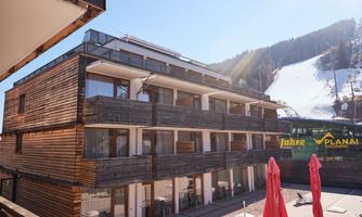 A modern hotel building with a wooden facade and large windows. In the background, snow-covered mountains and a ski resort can be seen.
