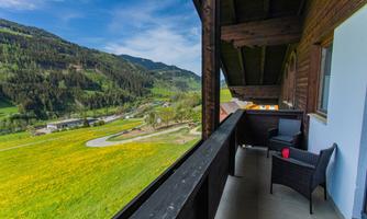 A balcony with a view of a green landscape and rolling mountains. In the corner, there are two chairs and a table, perfect for relaxing.
