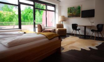 A bright room with two beds, a cozy armchair, and a table. Large windows offer a view of the green surroundings.
