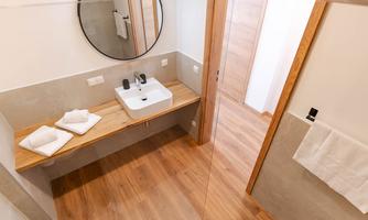 A modern bathroom with a large mirror and wooden shelf. The wall is light and the floor is made of wood.