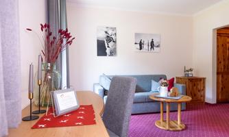 A cozy living room with a gray sofa and a wooden table.  
Pictures hang on the wall, and a vase with red plants is on the table.