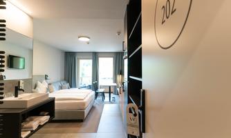 A modern hotel room with a large bed, a desk, and a window overlooking the outside. The walls are bright, and the room feels friendly and inviting.