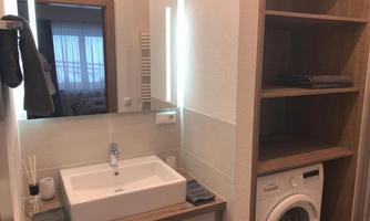 A modern bathroom with a sink and a large mirror. Next to the sink, there is a washing machine and wooden shelves.