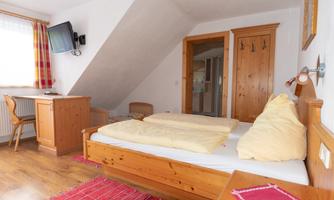 A bright bedroom with a double bed and a table. The room has wooden paneling and a television on the wall.
