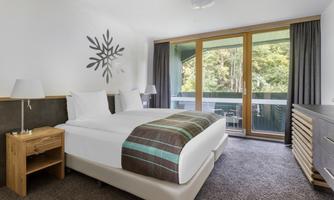 A modern hotel room with a cozy double bed and a desk. Large windows offer a view of a balcony and plenty of daylight.