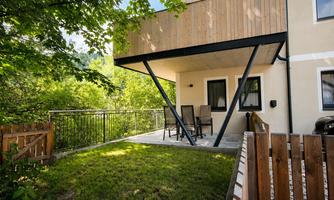 A modern terrace with a table and chairs, surrounded by green trees. The building has an elegant wooden facade and glass balustrade.