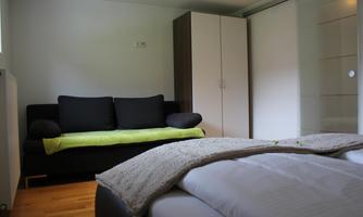 A cozy room with a black sofa and a bed. The room has a light wooden floor and a wardrobe.