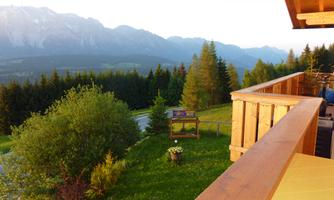 A picturesque view from a balcony overlooking green meadows and mountains. The image depicts a tranquil natural landscape with trees and a clear sky.