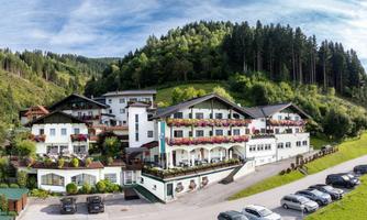 A charming hotel surrounded by green hills and trees. It offers a welcoming atmosphere with blooming balconies and a parking lot in the front.
